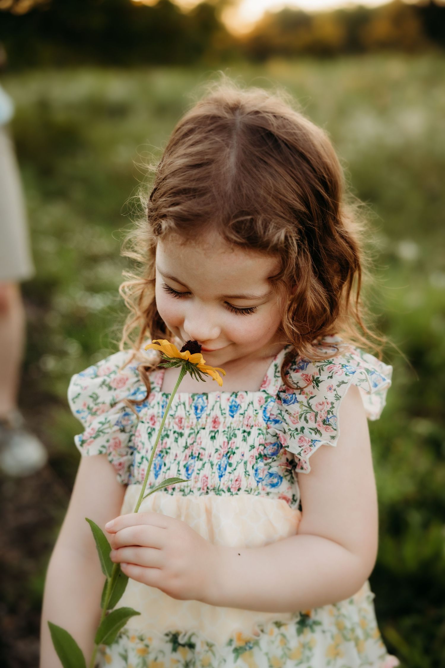Young girl in floral dress smelling a yellow black-eyed Susan wildflower in a sunny green meadow.