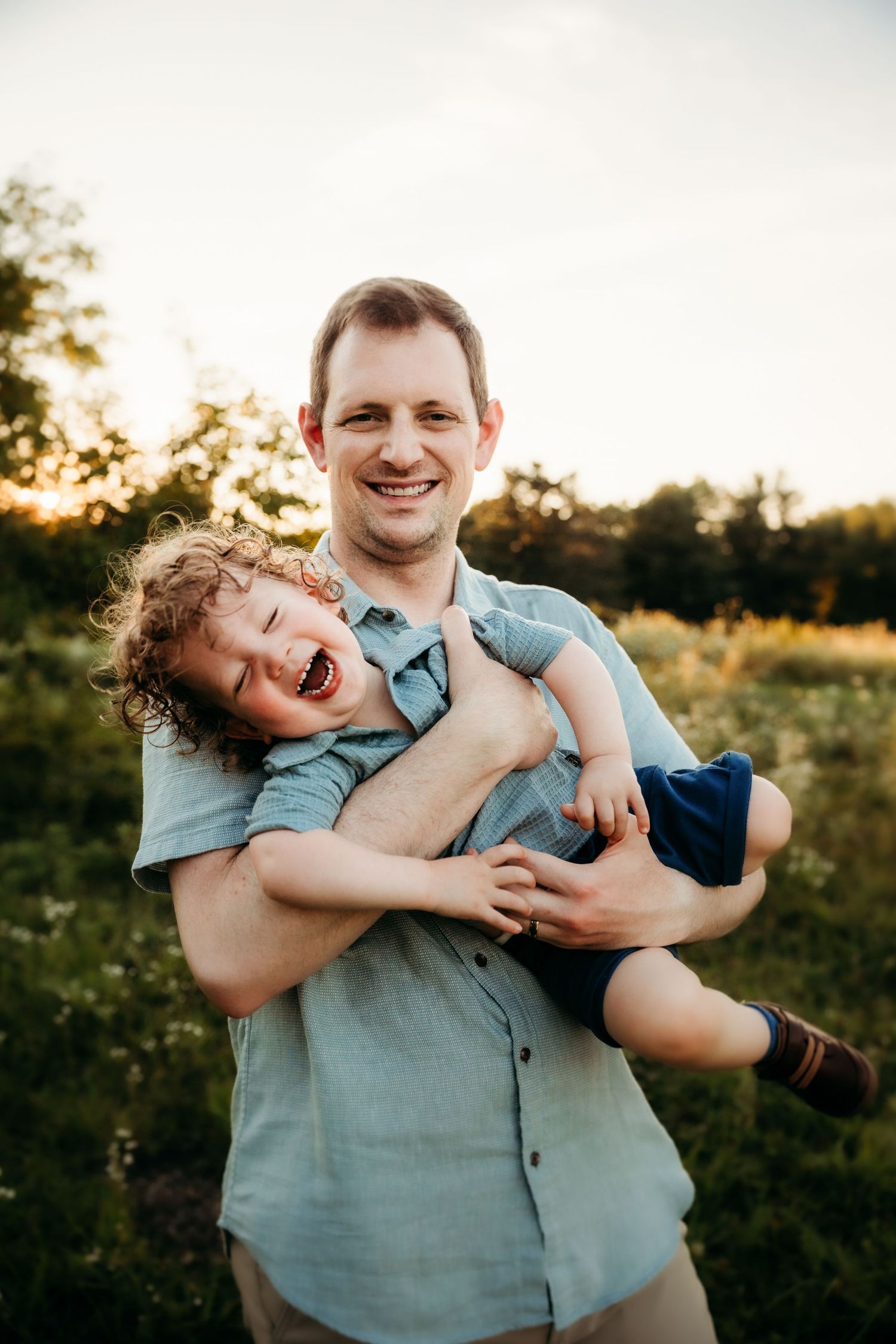 A smiling father holds his laughing curly-haired child outdoors at golden hour in a green field.