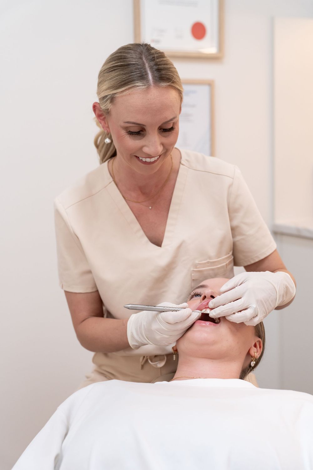 Smiling female dentist in beige scrubs and gloves performing a dental examination on a patient in a clinic.