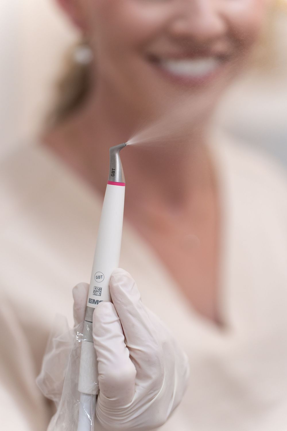 Dental professional in gloves holds a white teeth whitening pen emitting steam, with a smiling woman in the background.
