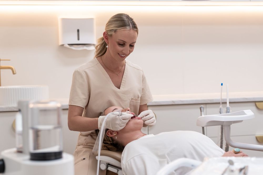 A dental hygienist in beige scrubs performs a teeth whitening treatment on a patient in a modern clinic.