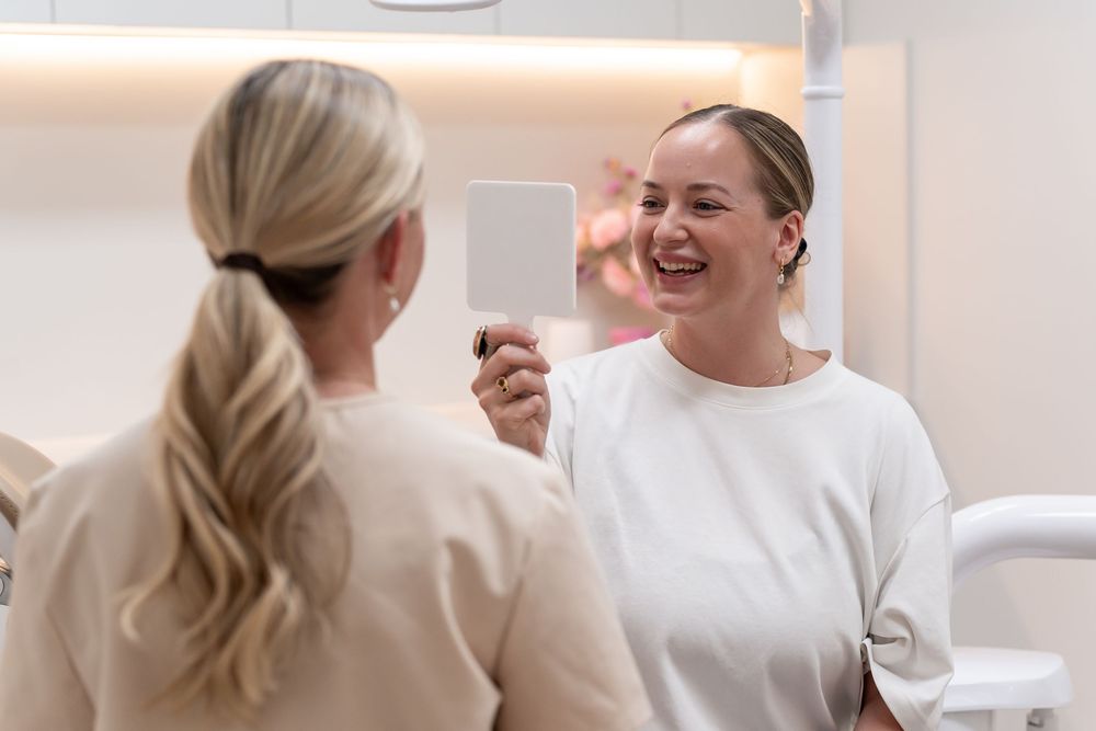A smiling woman examines her teeth in a handheld mirror at a dental clinic while a blonde professional looks on.