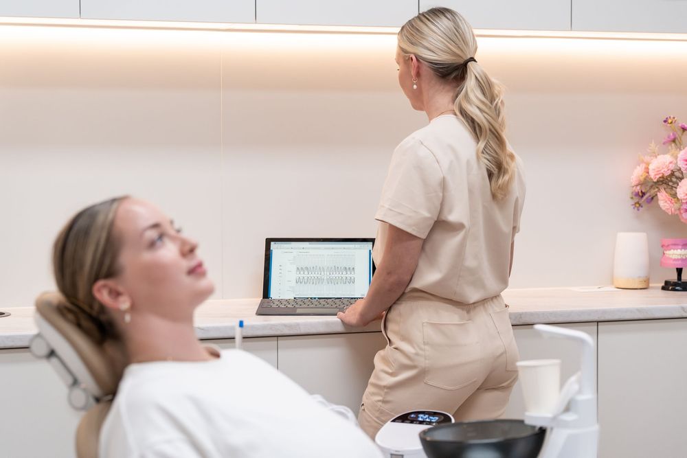A blonde aesthetician reviews treatment notes on a laptop while a relaxed female client reclines in a modern clinic chair.