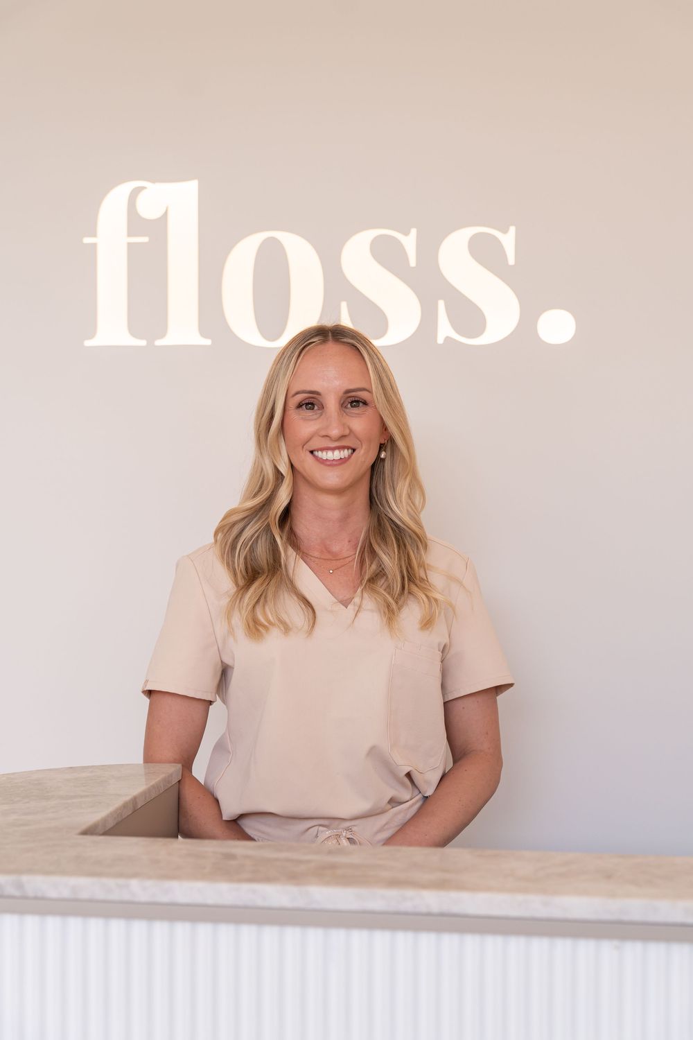 Smiling blonde woman in beige scrubs standing at a dental reception desk with Floss logo on wall.