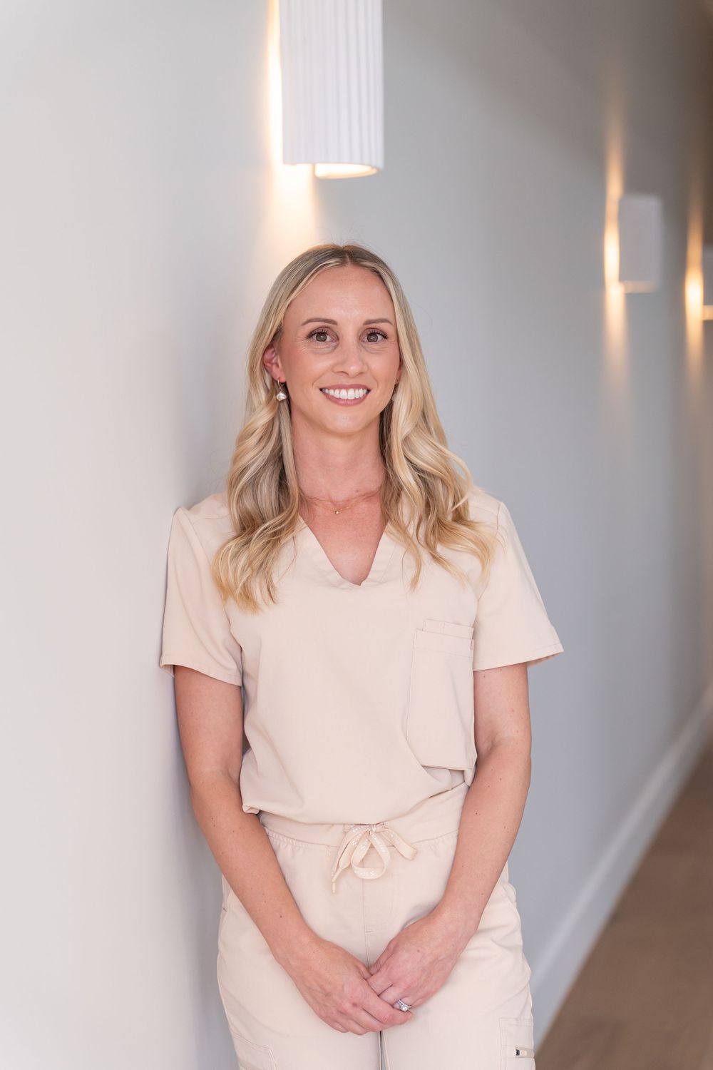 A smiling female dental professional in beige scrubs stands in a bright modern clinic hallway.
