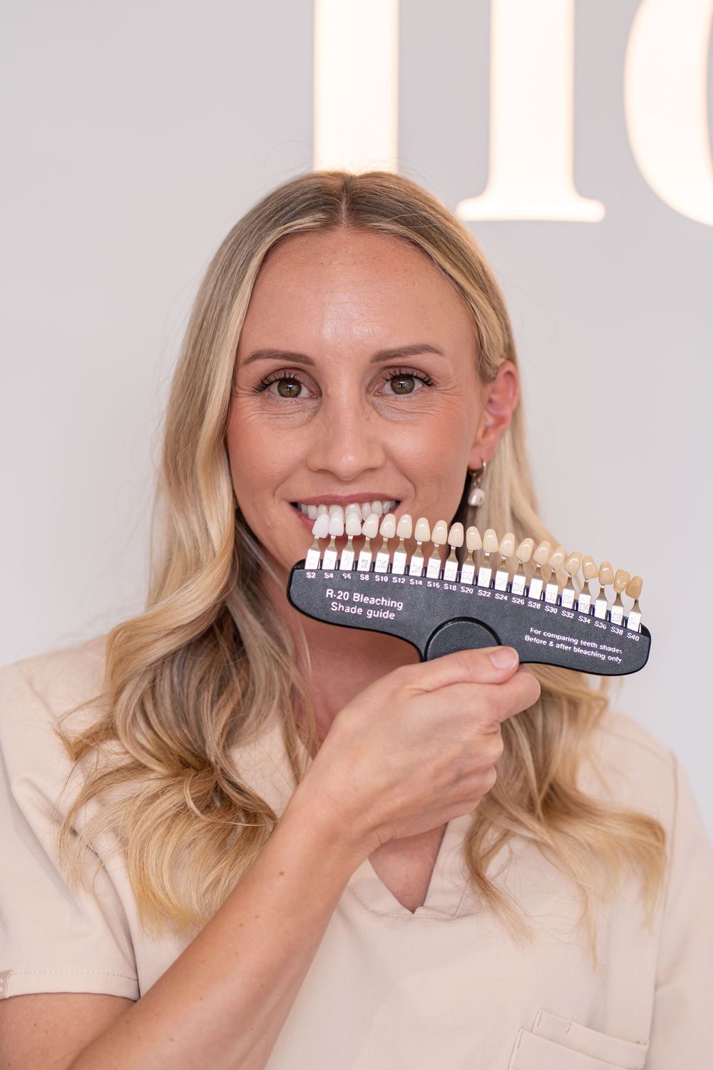 Smiling blonde woman holds a dental shade guide next to her teeth for a whitening comparison at a dental clinic.