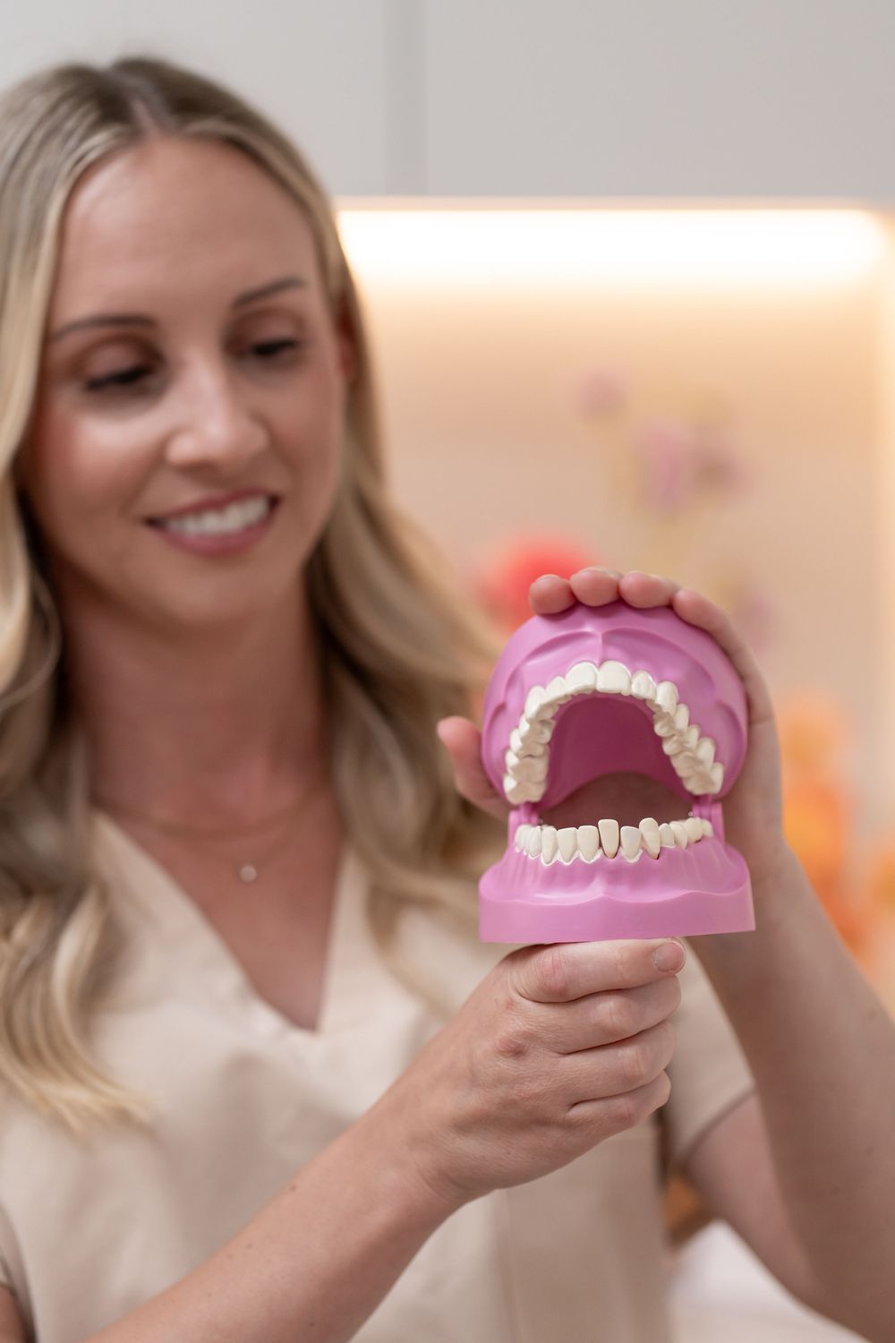 Smiling blonde dentist holding a pink dental jaw model demonstrating teeth in a modern dental office.