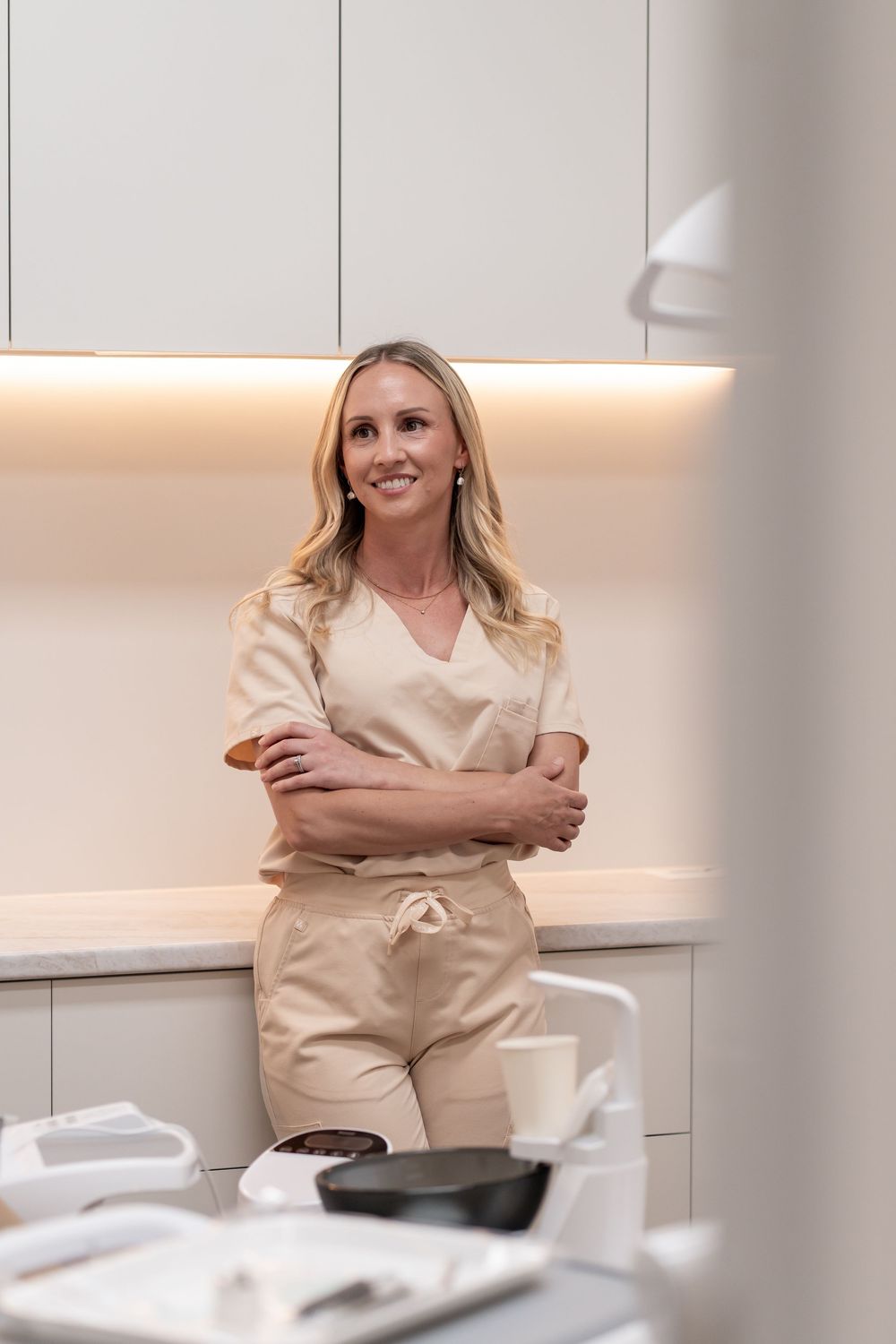 Smiling female dentist in beige scrubs standing with arms crossed in a modern dental clinic.