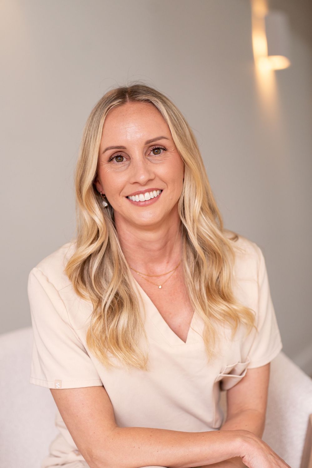Smiling blonde woman in a cream v-neck top poses for a professional headshot in a softly lit studio.