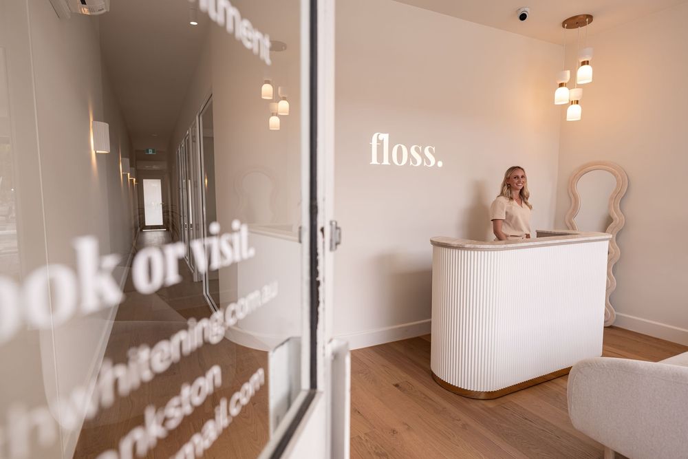 Modern dental clinic reception area with white decor, gold accents, and a receptionist at the Floss whitening studio desk.