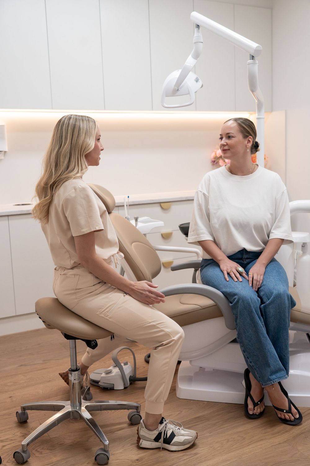 A dentist in beige scrubs consults with a patient seated in a dental chair in a modern clinic.