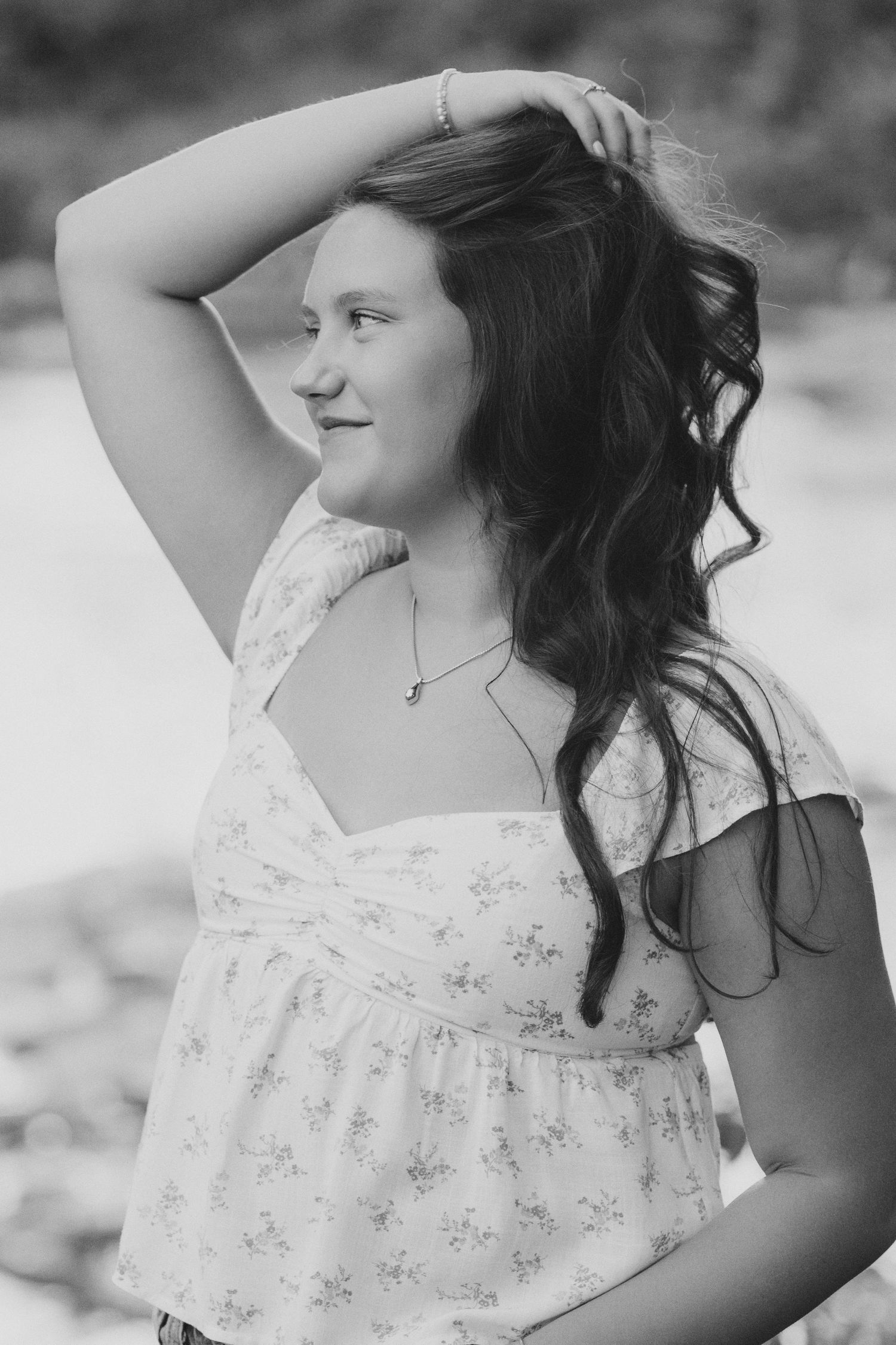Black and white portrait of a young woman in a floral dress, hand in hair, smiling outdoors.