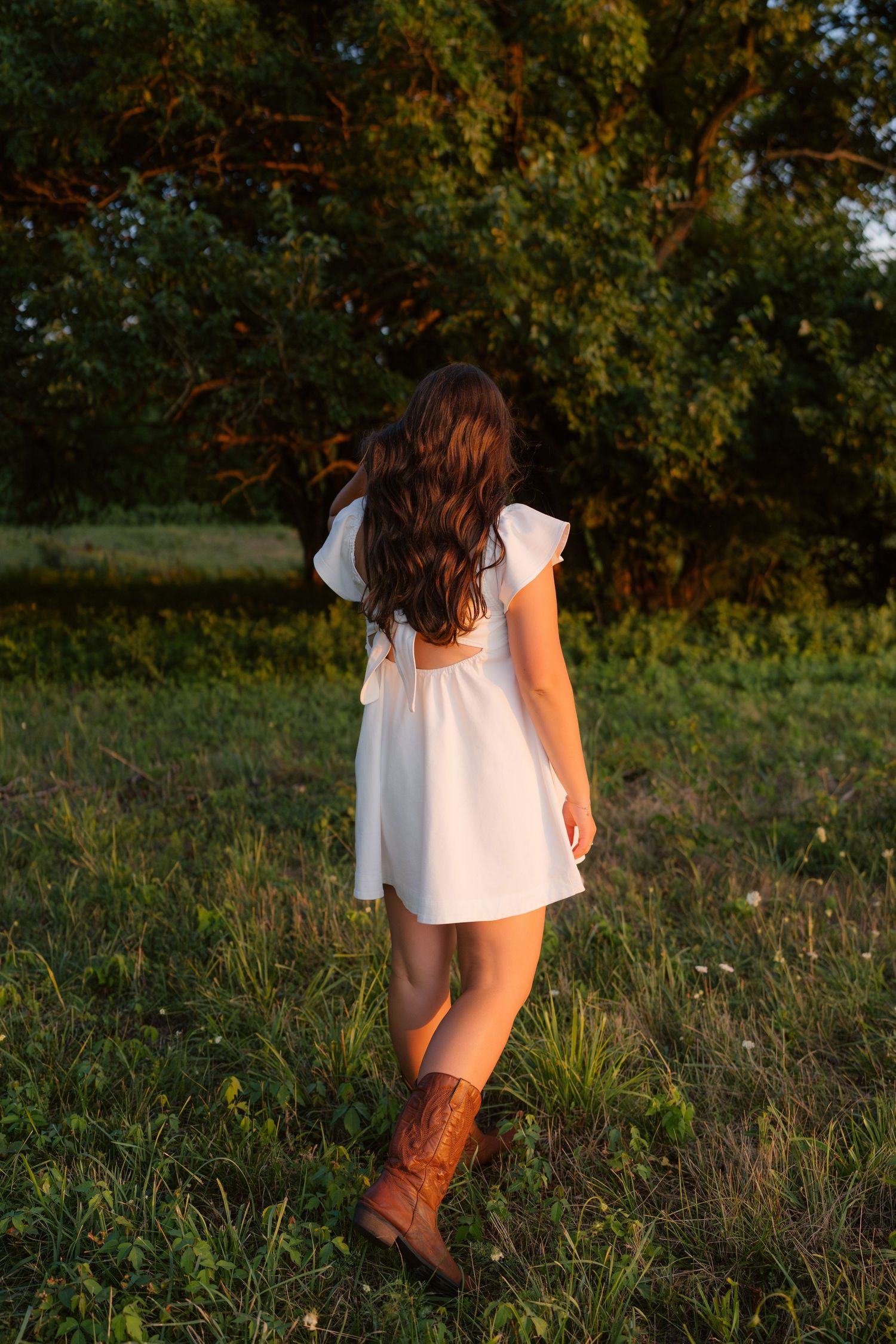 Young woman in white dress and brown cowboy boots walking through green meadow at golden hour sunset.