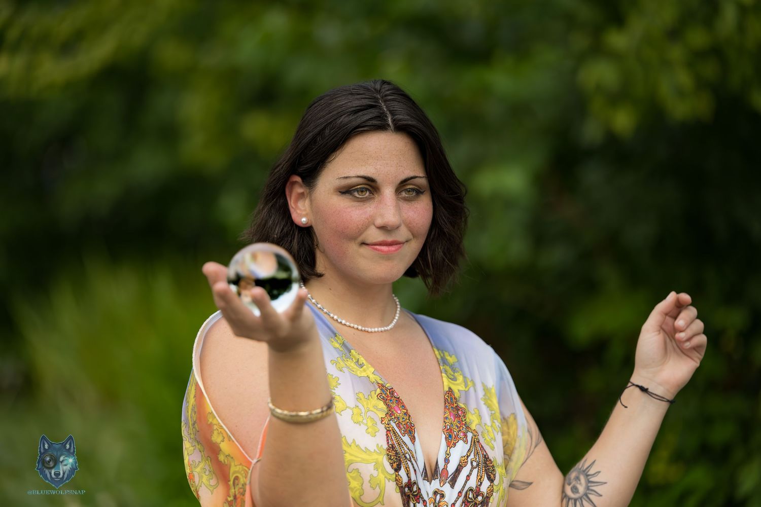 Young woman in colorful top holds small crystal ball in lush green outdoor setting. Chi.Shooters meet gabbsbosh
