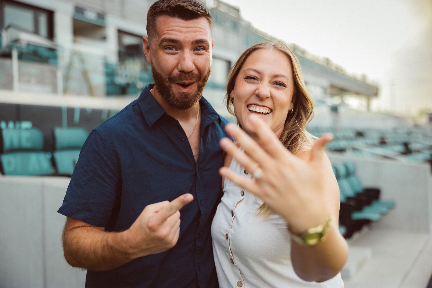 A woman holds her new engagement ring up to the camera as her fiancé points to it proudly after pulling off a surprise stadium proposal.