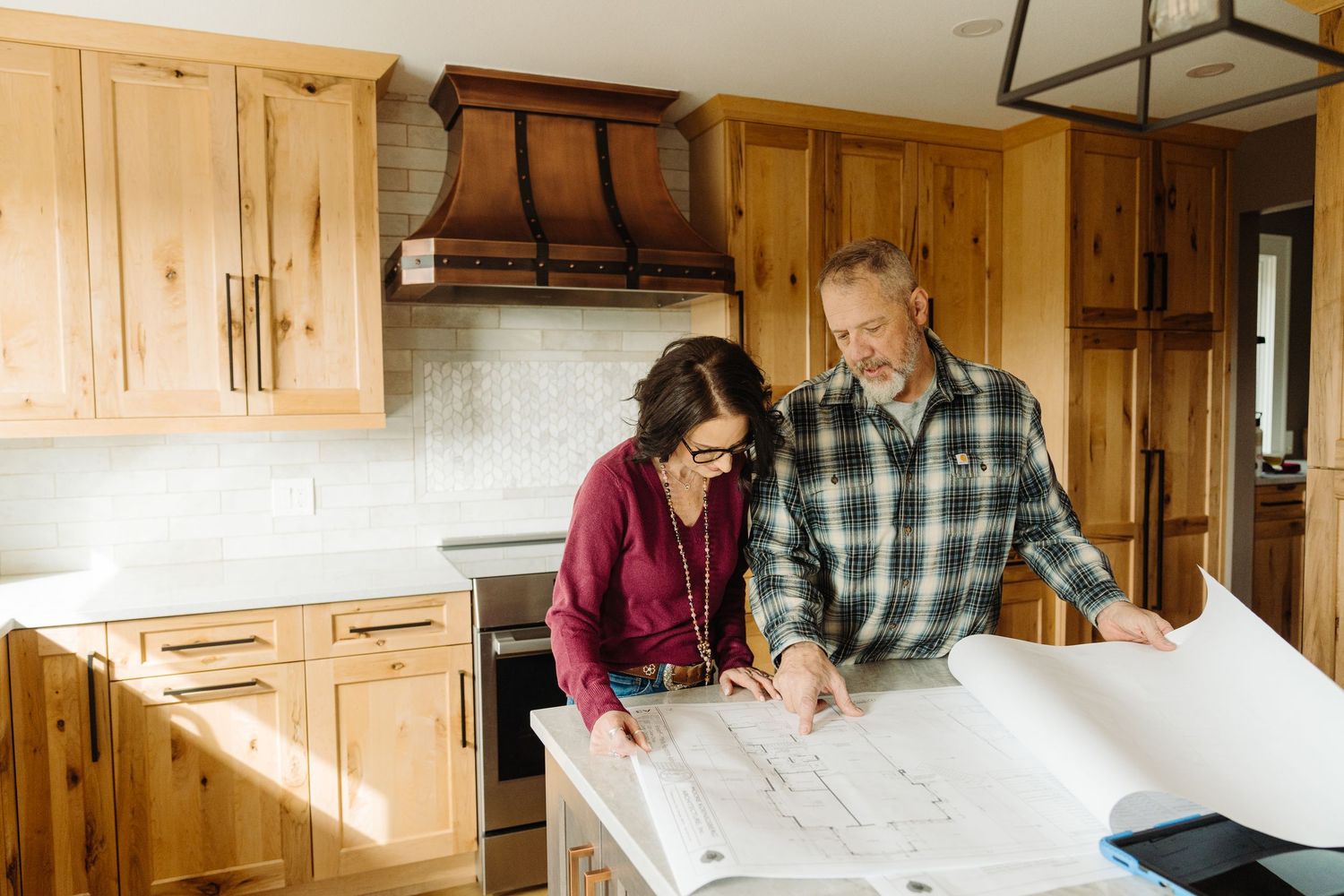 Couple reviewing architectural blueprints together in a rustic wooden kitchen with natural cabinetry and tile backsplash.