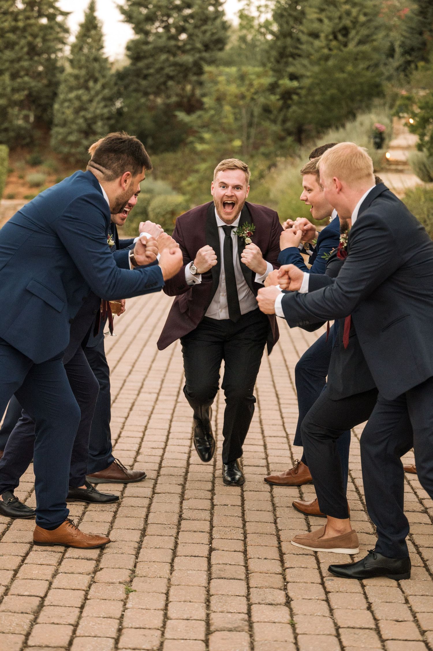 Groom in burgundy suit jumps with excitement surrounded by cheering groomsmen in navy suits on a brick pathway.