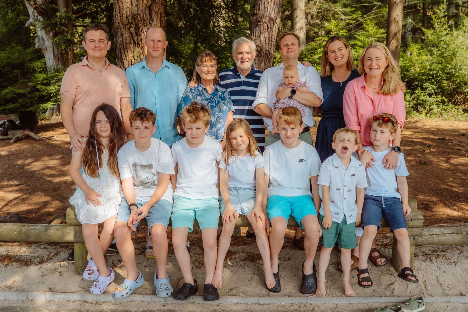 Large multigenerational family group photo with adults and children posing outdoors among tall trees on a sunny day.