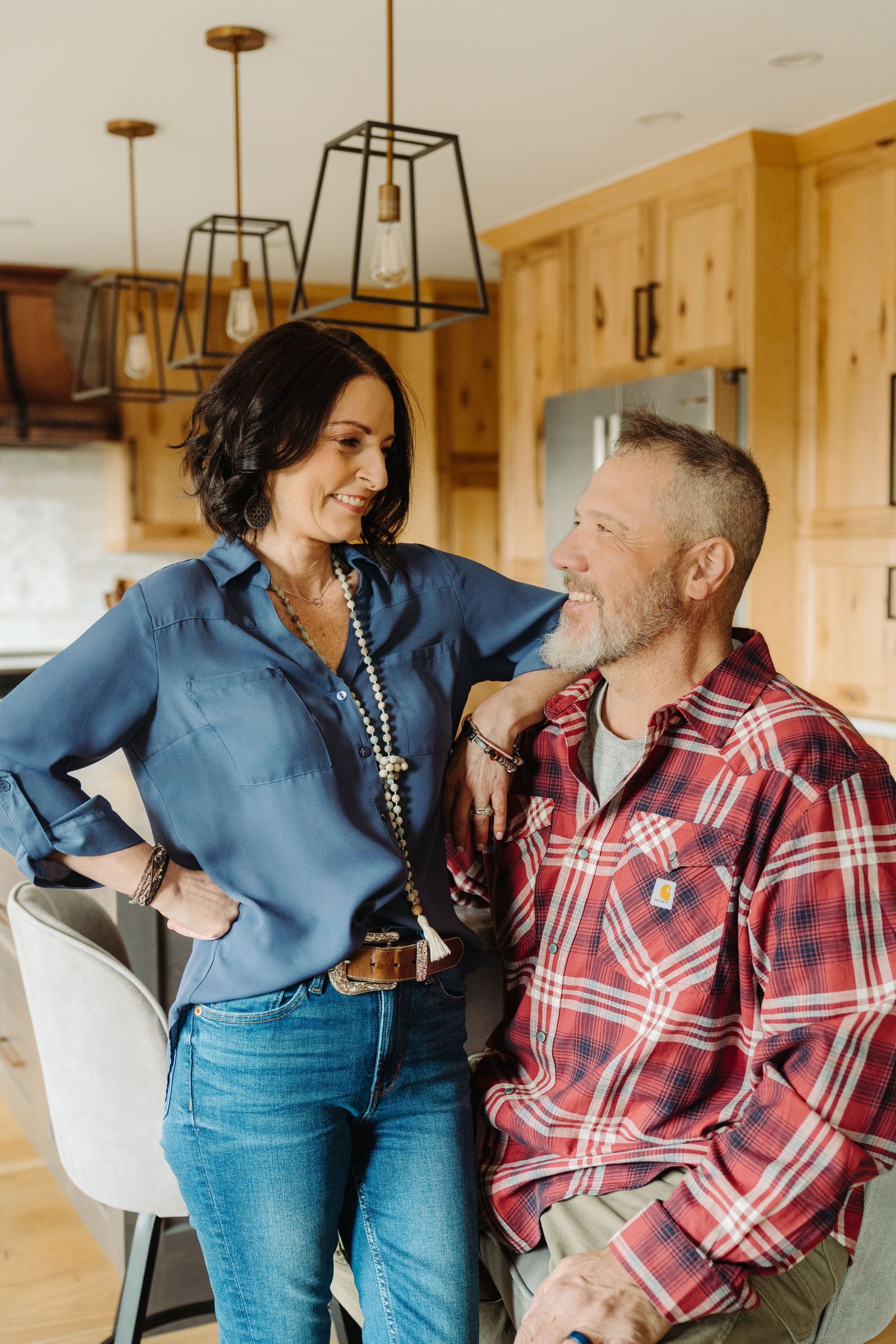 A smiling middle-aged couple in casual attire share a warm moment together in a modern rustic kitchen.