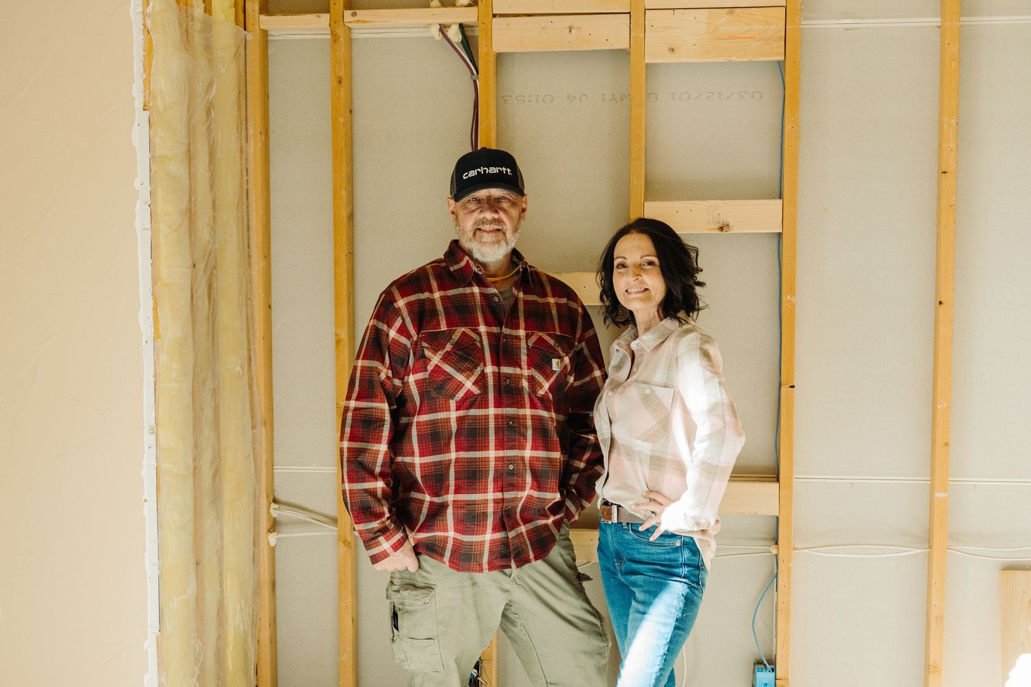 A man and woman smiling together inside a home under renovation with exposed wooden wall framing.