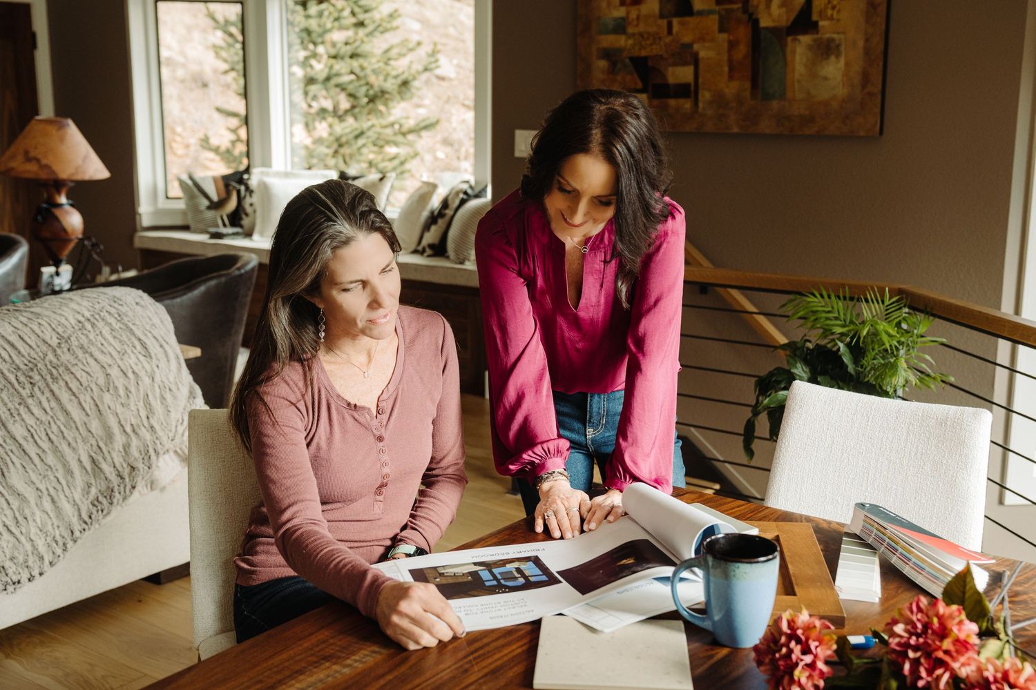 Two women reviewing documents together at a home office desk with a laptop, coffee, and decorative flowers.