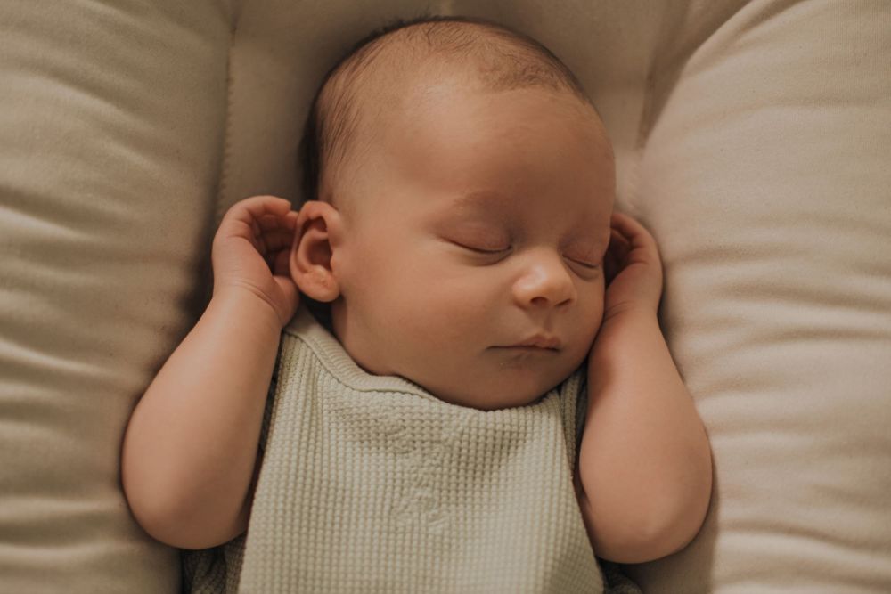 Peaceful newborn baby sleeping soundly in a soft beige nest, wearing a light green waffle onesie with tiny hands raised.