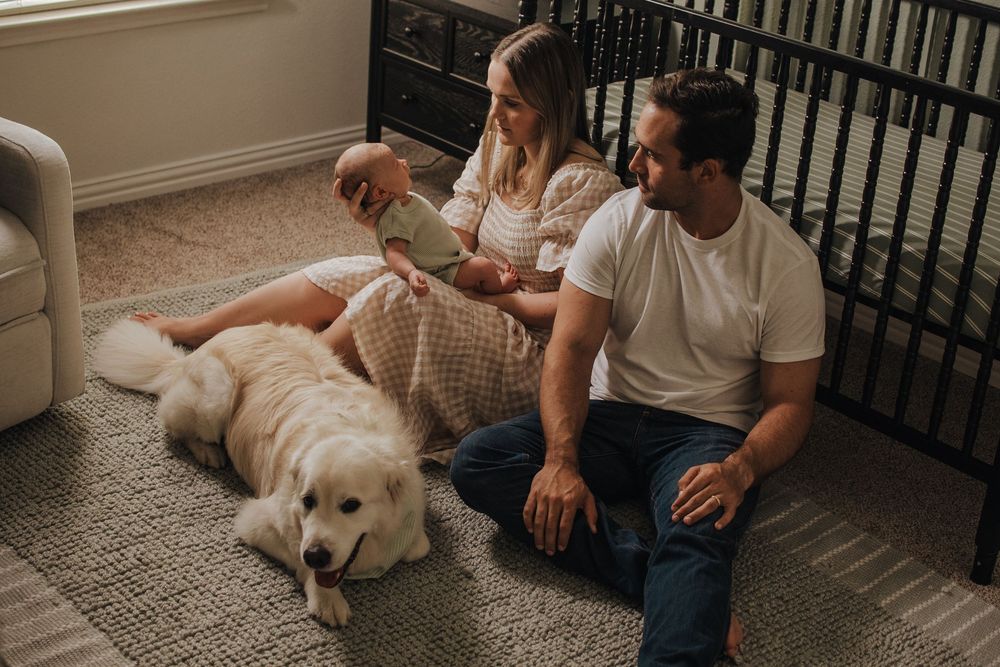 Young couple sitting on nursery floor with newborn baby and golden retriever dog near crib.