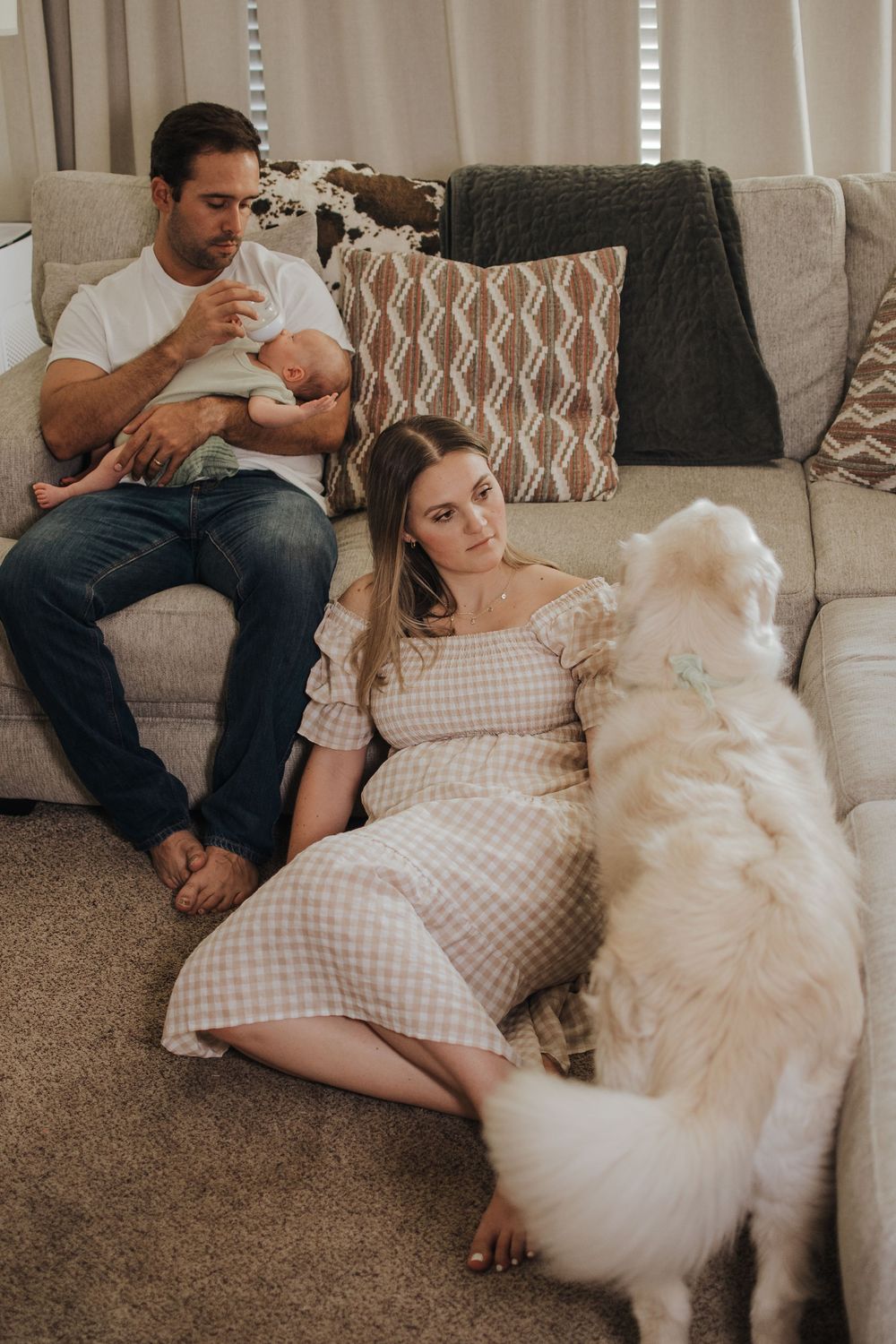 Family with newborn baby and white dog relaxing on living room couch and floor in cozy home setting.