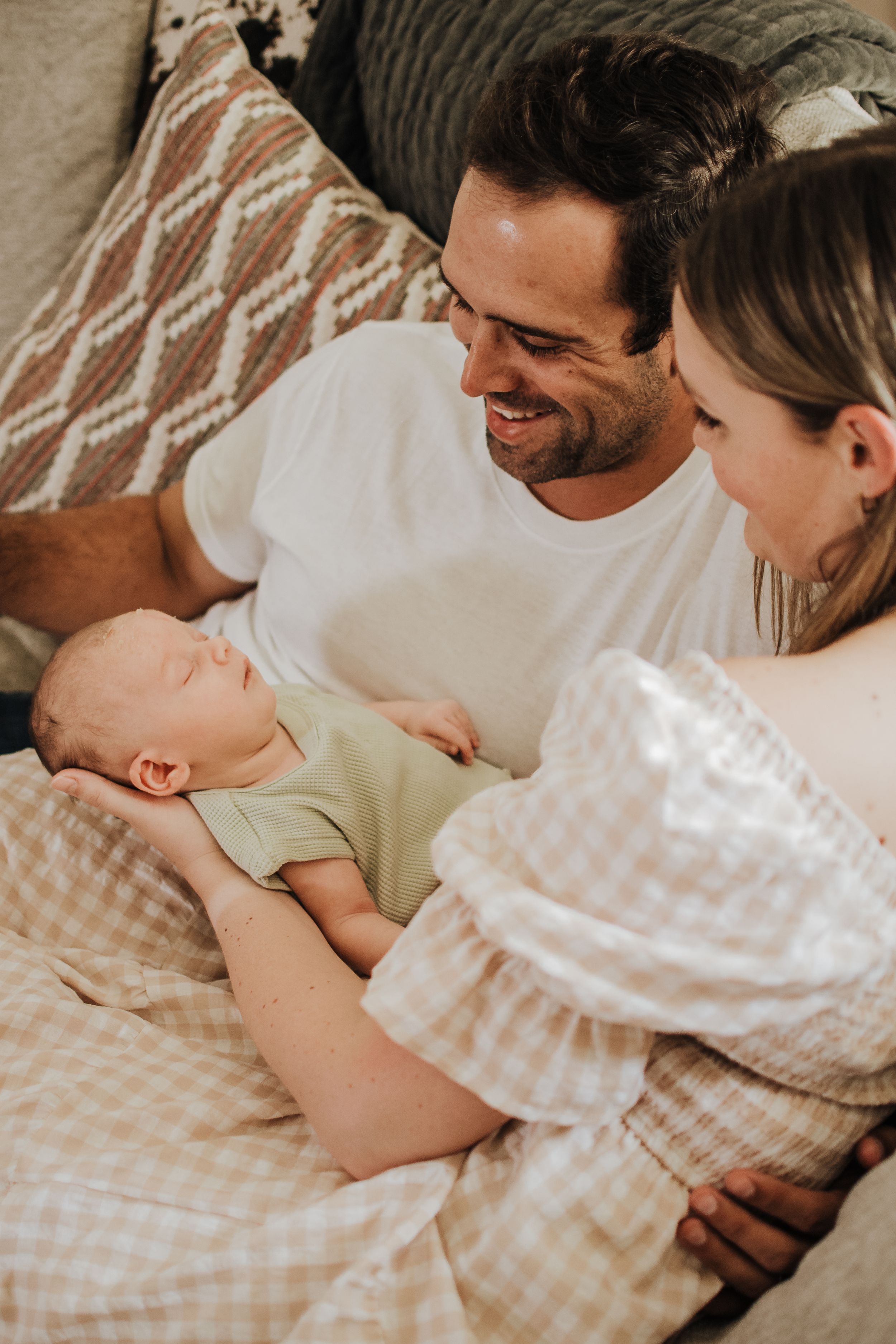 Happy parents smiling at their sleeping newborn baby lying on a gingham blanket between them.