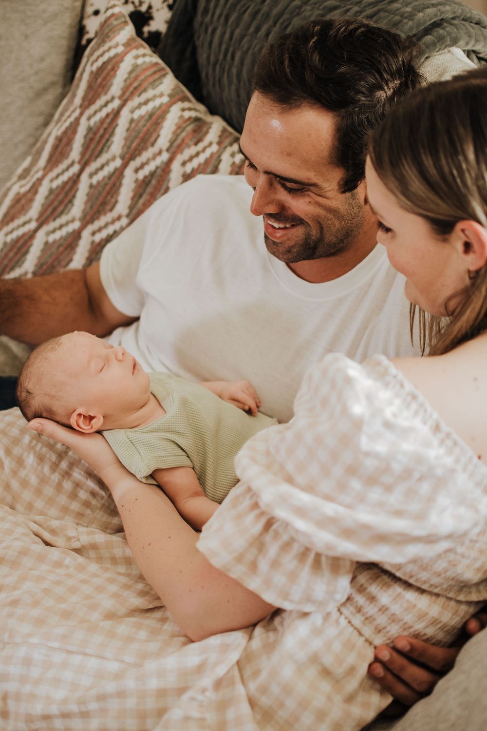 Happy parents smiling at their sleeping newborn baby lying on a gingham blanket between them.