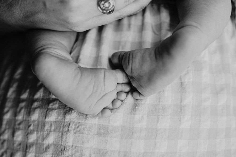 Black and white photo of a newborn baby's tiny feet held gently by an adult hand on a soft blanket.