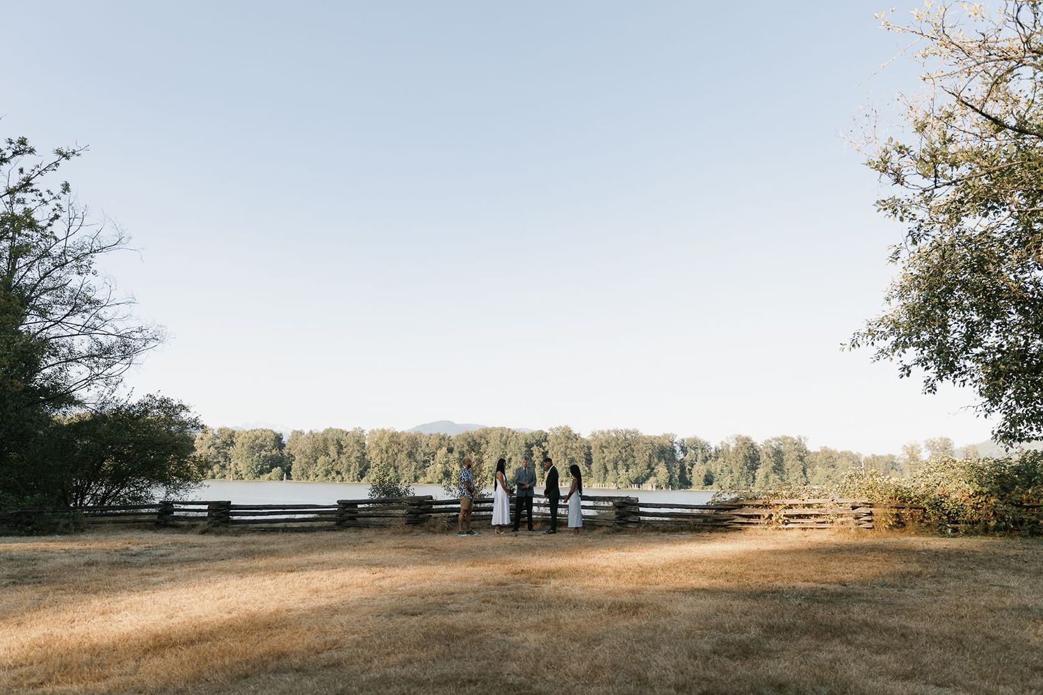 Wedding party gathered near a rustic fence by a serene lake, surrounded by lush trees under a clear blue sky.