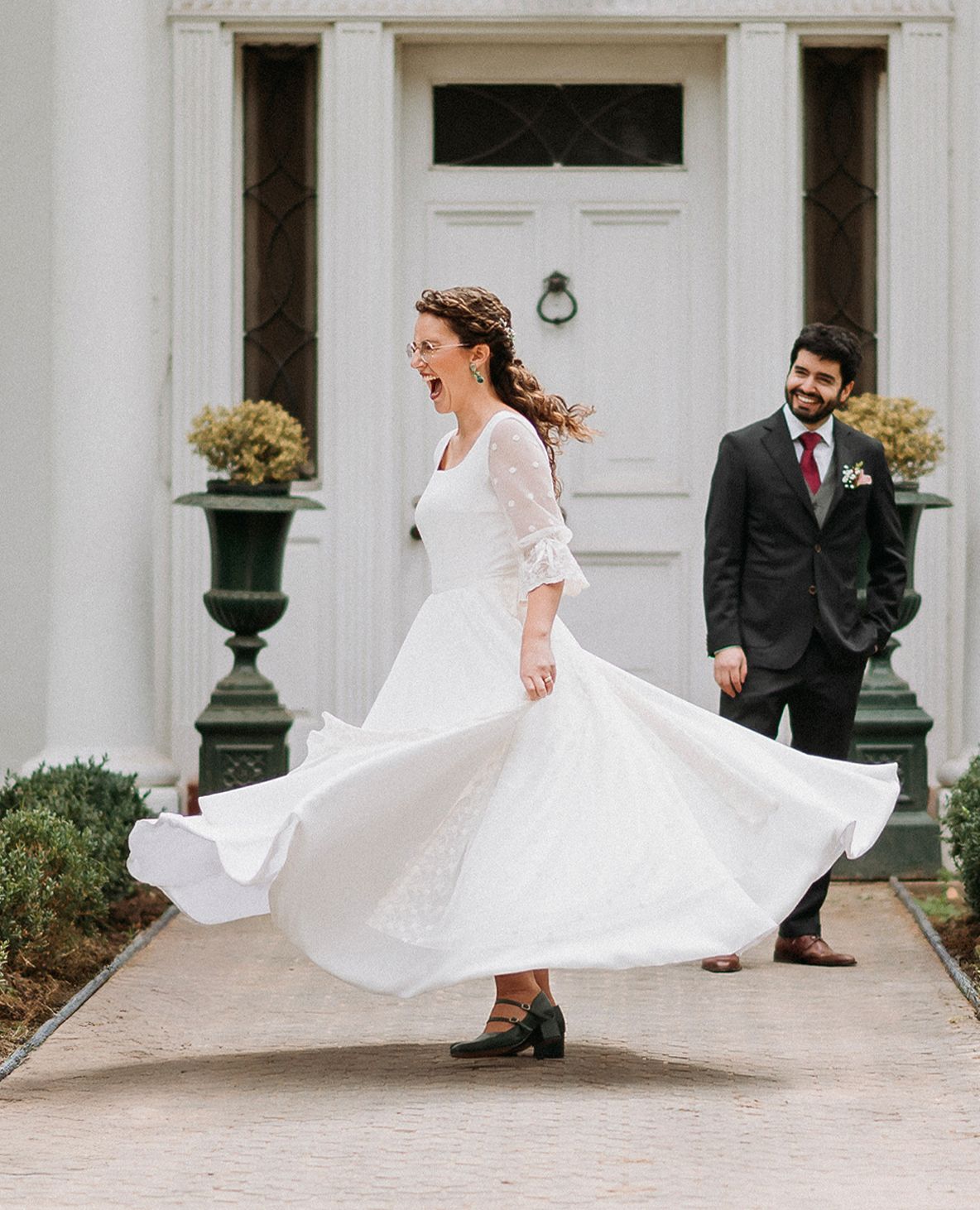 Bride twirling white dress outside white manor as groom watches.