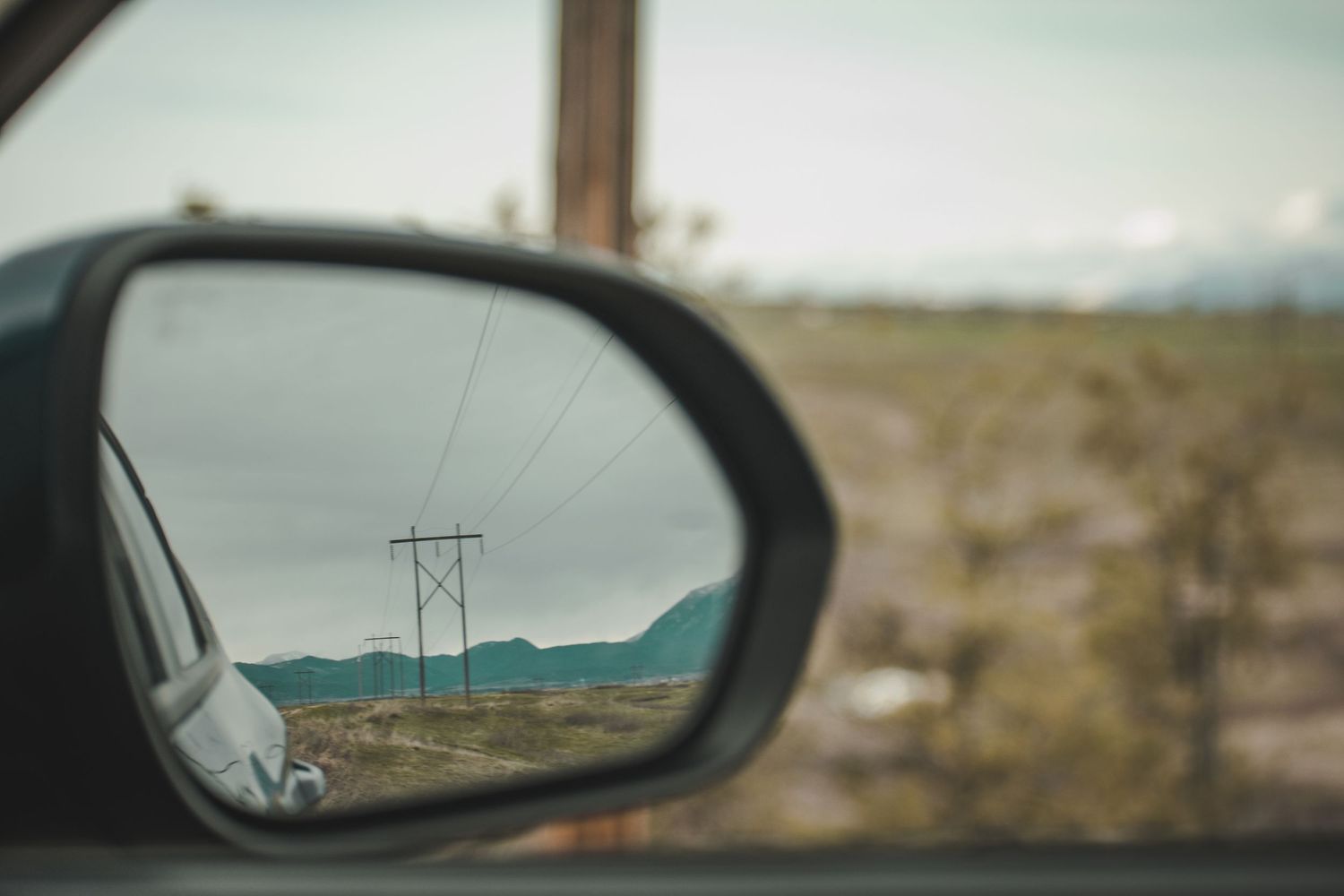 Car side mirror reflecting power lines, mountains, and open desert landscape on a road trip.
