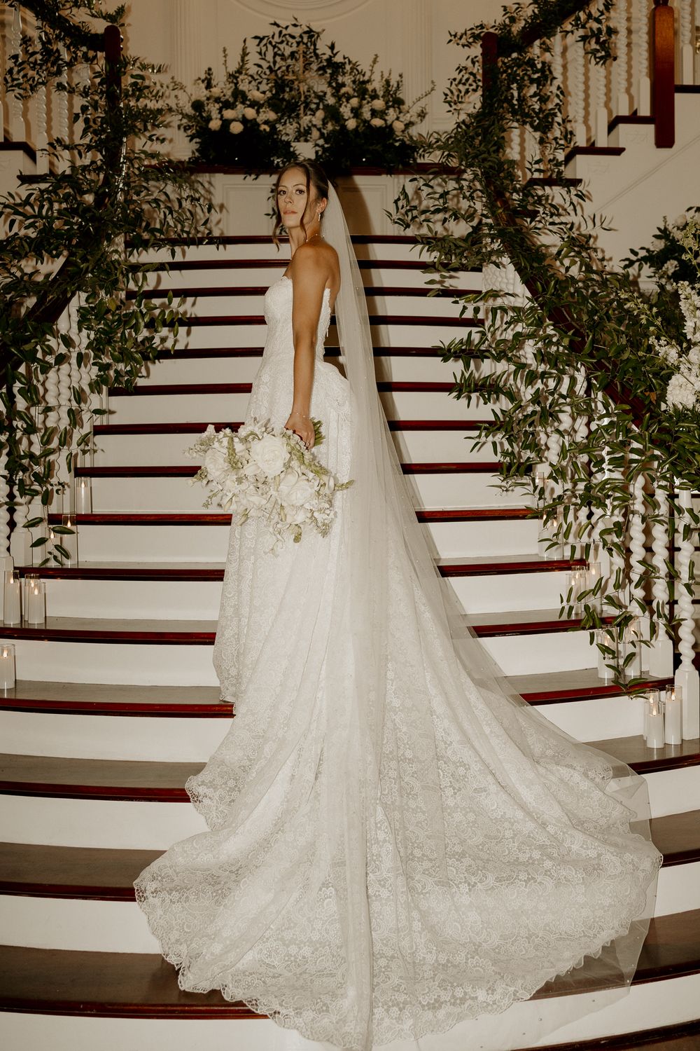 Bride in lace gown with long cathedral veil holding white bouquet on floral-decorated grand staircase.
