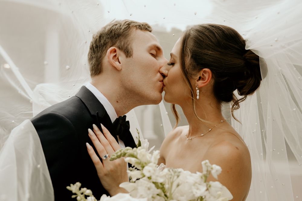 Bride and groom share a romantic kiss under a flowing veil, bride holding a white floral bouquet.