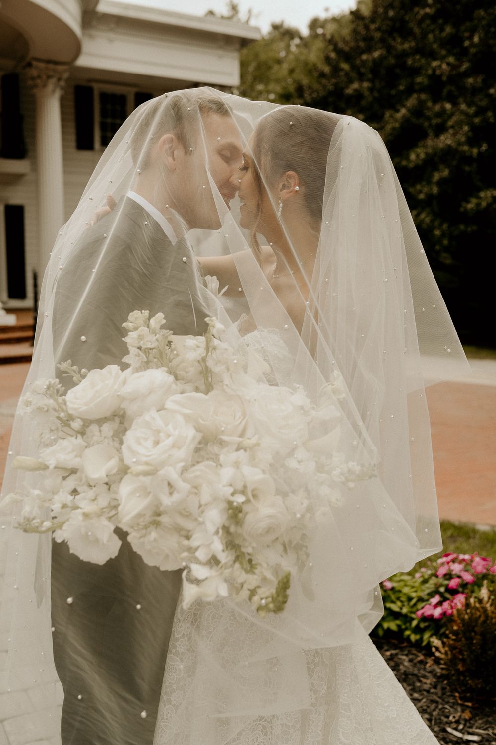 Bride and groom sharing intimate kiss under flowing veil holding white floral bouquet outdoors.