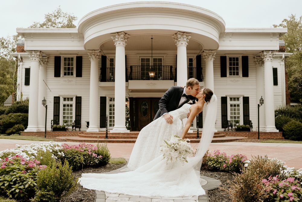 Bride and groom share a romantic dip kiss in front of a grand white Southern mansion with columns.