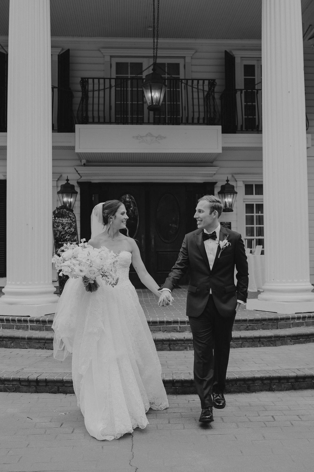 Black and white photo of bride and groom holding hands walking in front of elegant colonial venue.