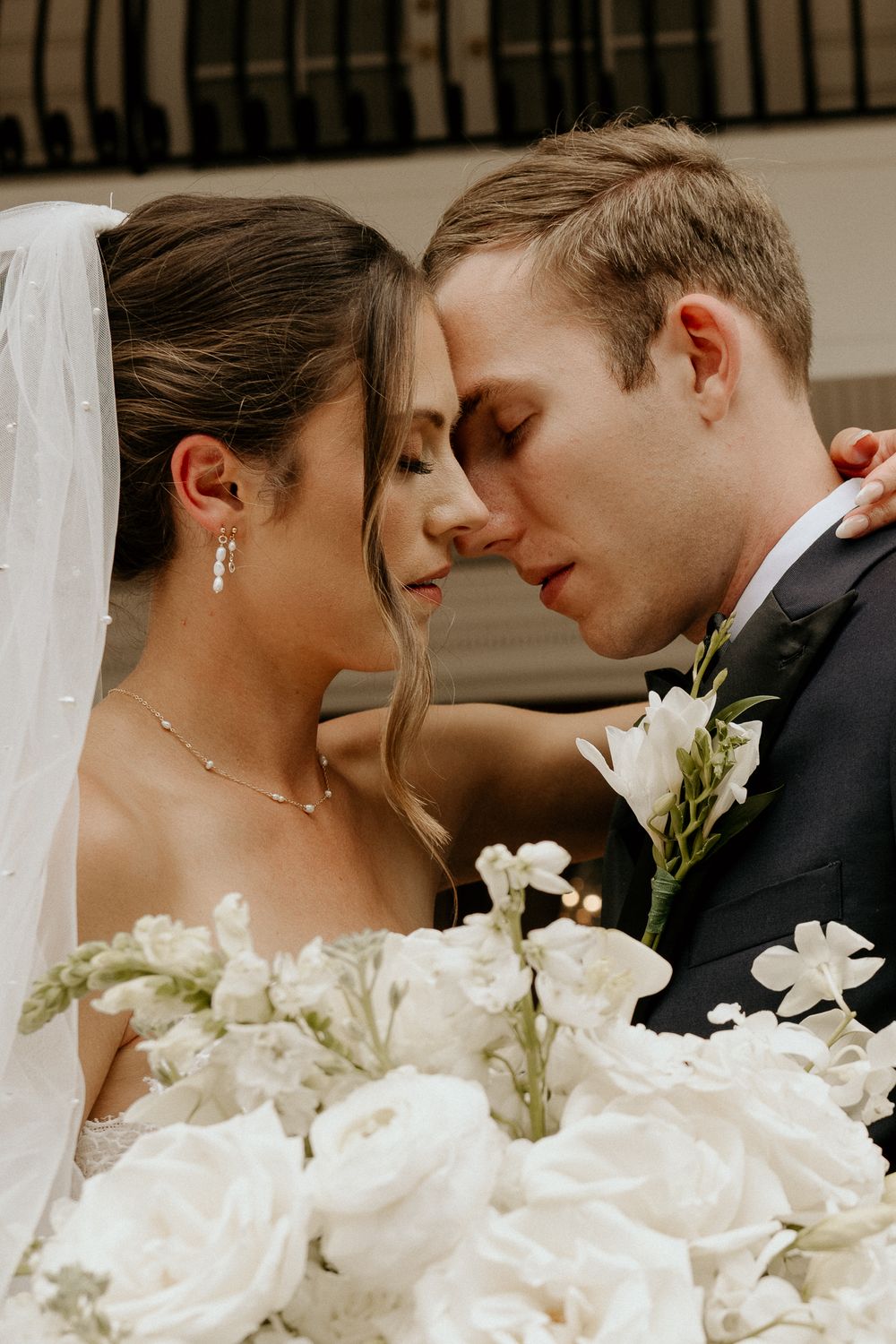 Close-up of bride and groom touching foreheads tenderly, bride holding lush white floral bouquet in elegant wedding attire.