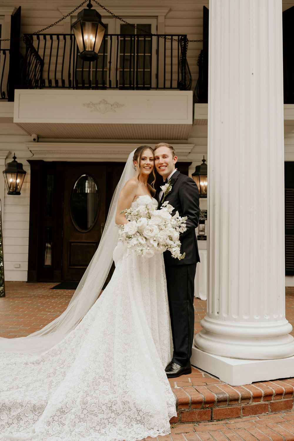 Bride and groom pose by white column, bride in lace gown with cathedral veil holding white bouquet.