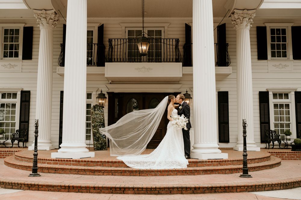 Bride and groom kissing on brick steps of white colonial mansion with dramatic veil flowing.