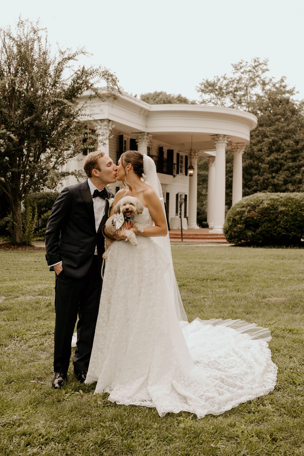 Bride and groom kiss on lush lawn holding a small dog near a white columned gazebo.