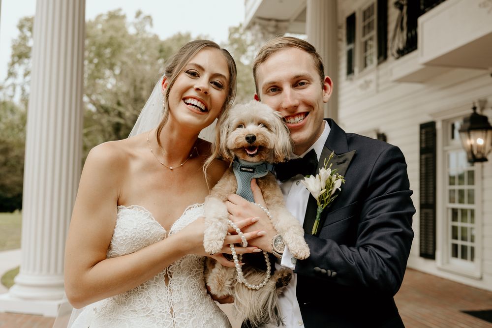 Smiling bride and groom pose with their adorable dog wearing a bow tie on wedding day.