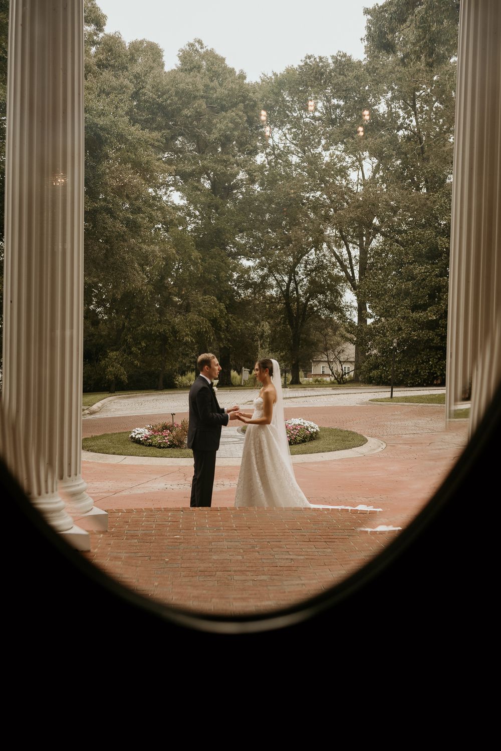 Bride and groom exchange vows outdoors between classic columns, framed by an oval window with lush green trees behind.