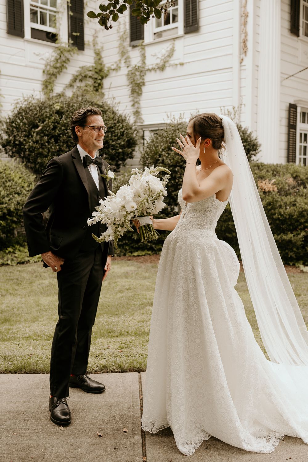Emotional bride covers face during first look with groom in black tuxedo outside white estate.