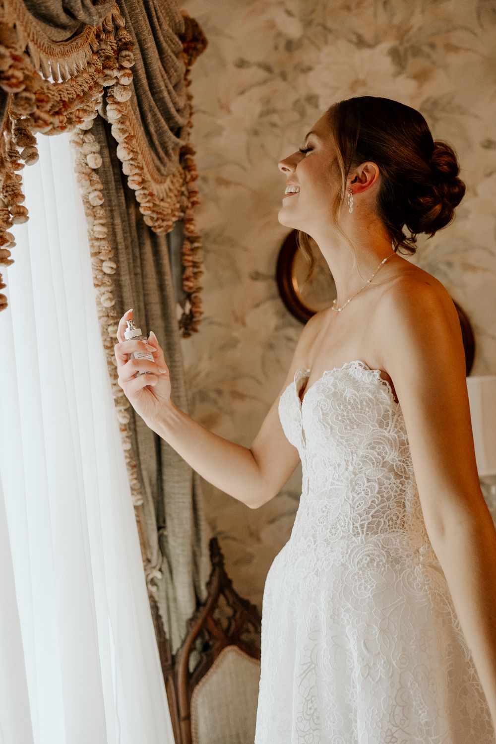 Smiling bride in strapless lace wedding gown applying perfume by a draped window in an elegantly wallpapered room.