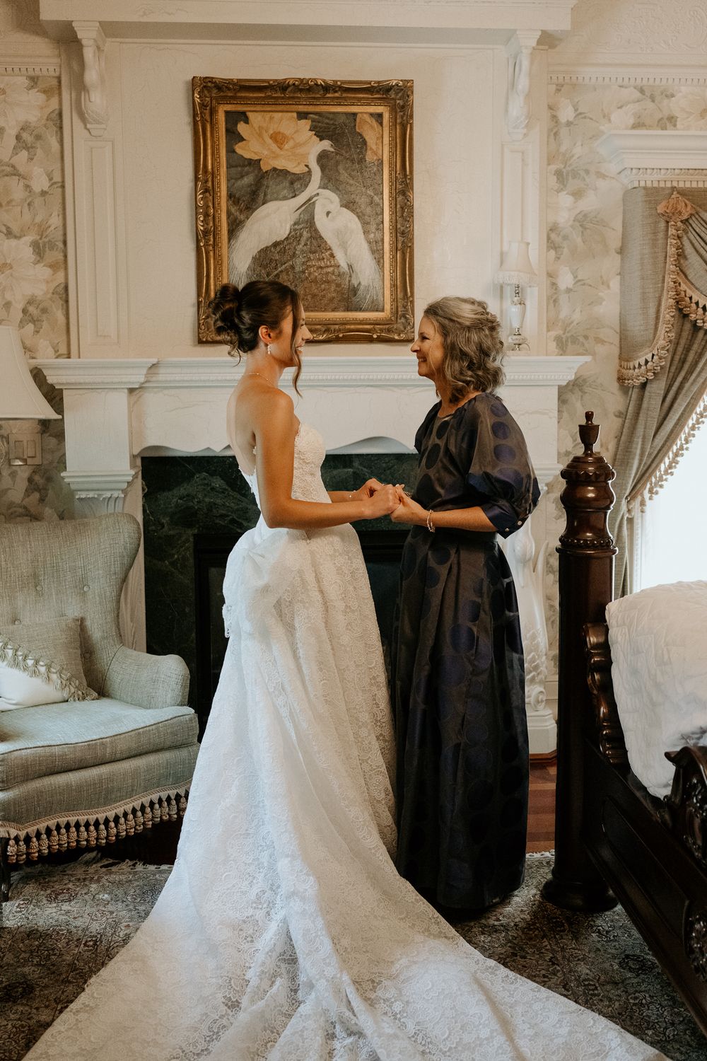 Bride in lace gown sharing tender moment with mother near ornate fireplace in elegant room.