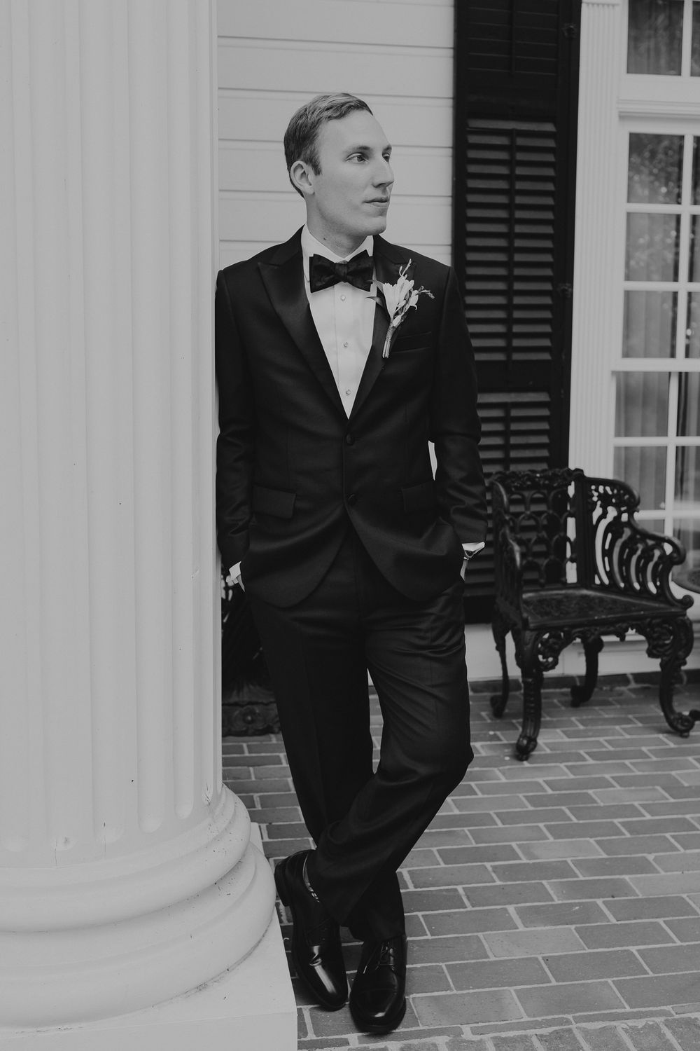 Dapper groom in black tuxedo with boutonniere leaning against white column in black and white photo.