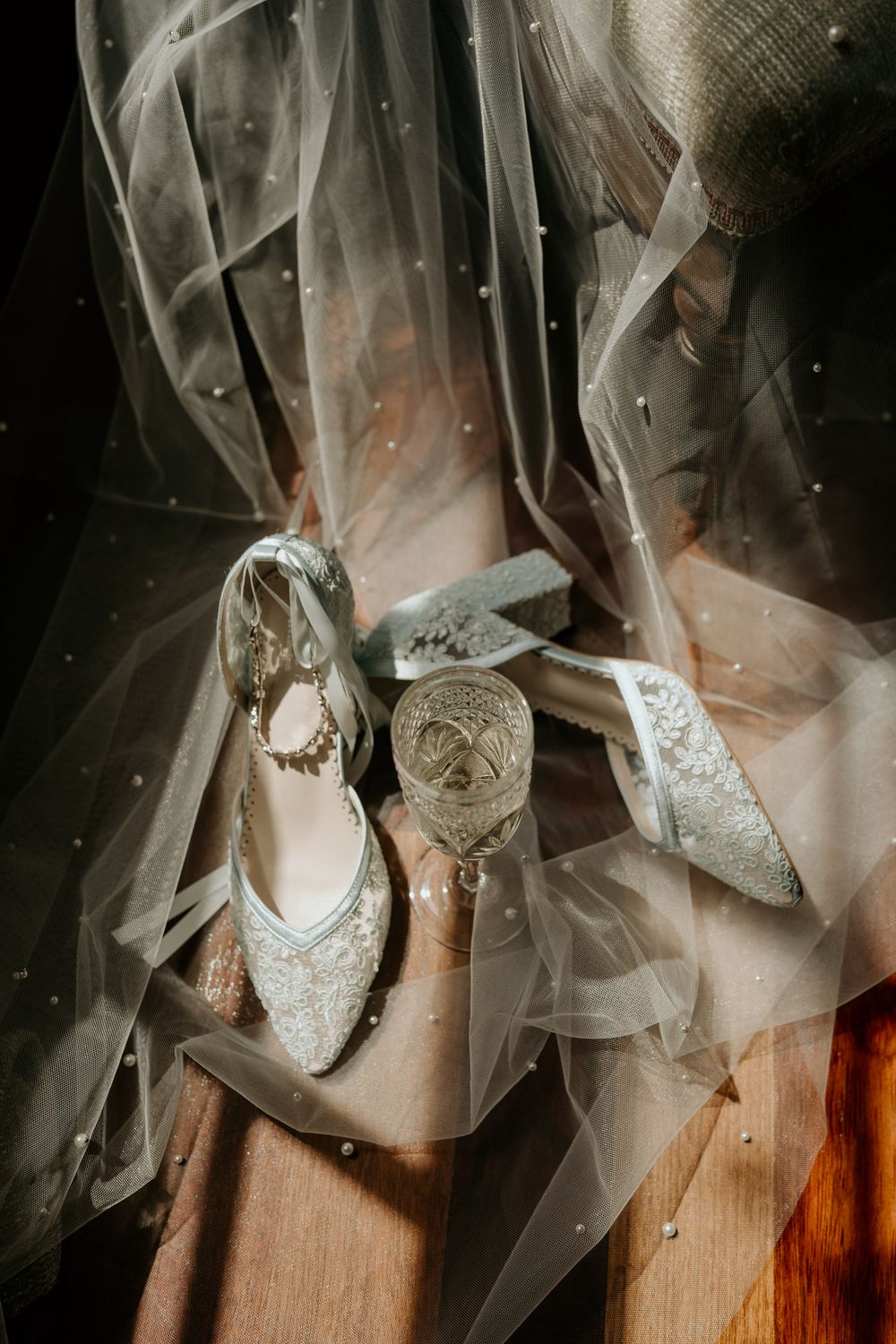 Elegant bridal heels with pearl embellishments and lace details displayed on wooden floor with sheer dotted veil.